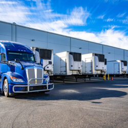 Blue big rig semi truck with extended cab for truck driver rest and refrigerator semi trailer standing in row with another semi trailers in warehouse dock gates loading cargo for next freight